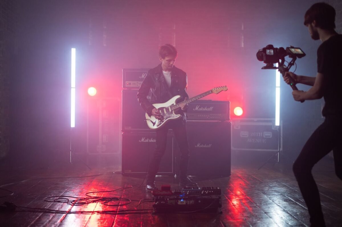 Guitarist in a leather jacket performs in a studio setting with vibrant lighting, captured by a cameraman.